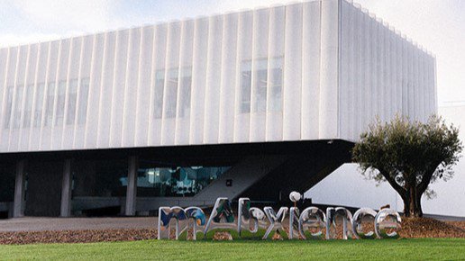 Modern white building with large windows and "mAbxience" sign in front, next to a tree.