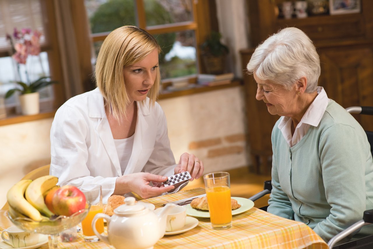 Nurse showing medicines to senior woman who is sitting in a wheelchair at dining table.