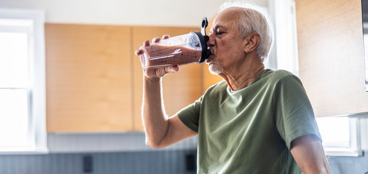 An older adult drinks a blended nutritional beverage while seated indoors.