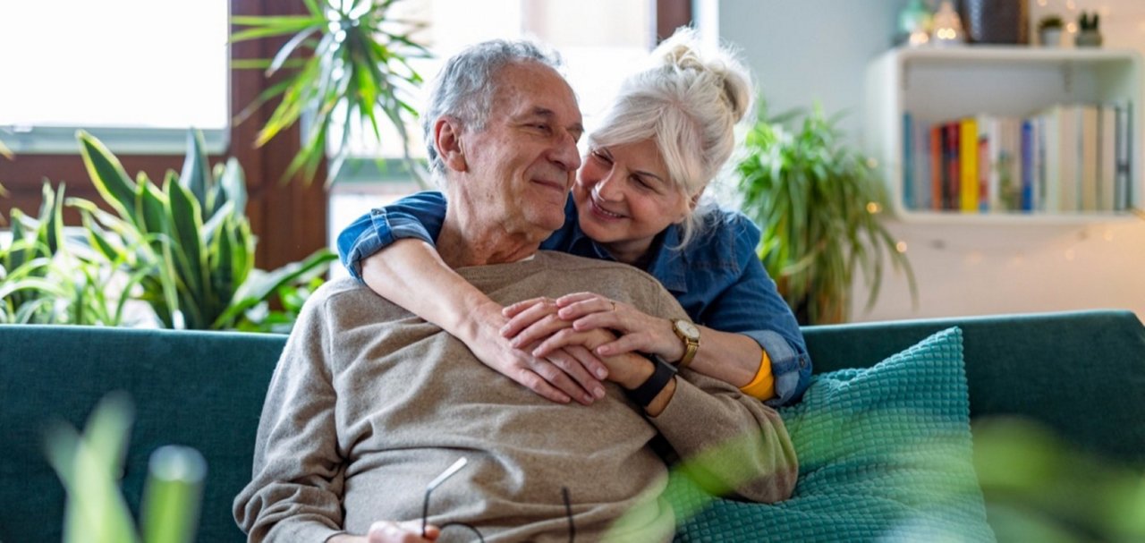 Two older people sitting closely together on a sofa in a plant-filled living room