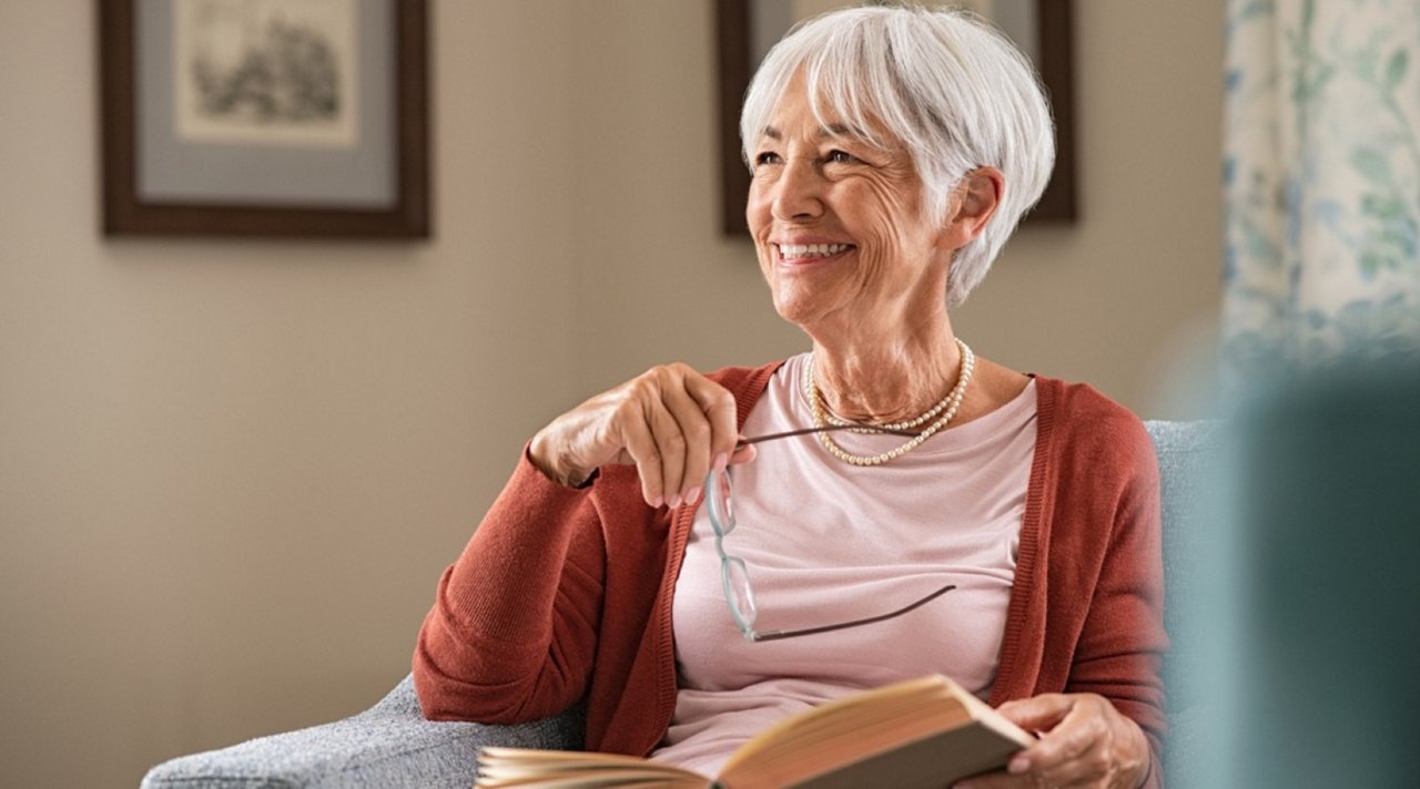 Older person sitting in an armchair with an open book and holding a pair of glasses.