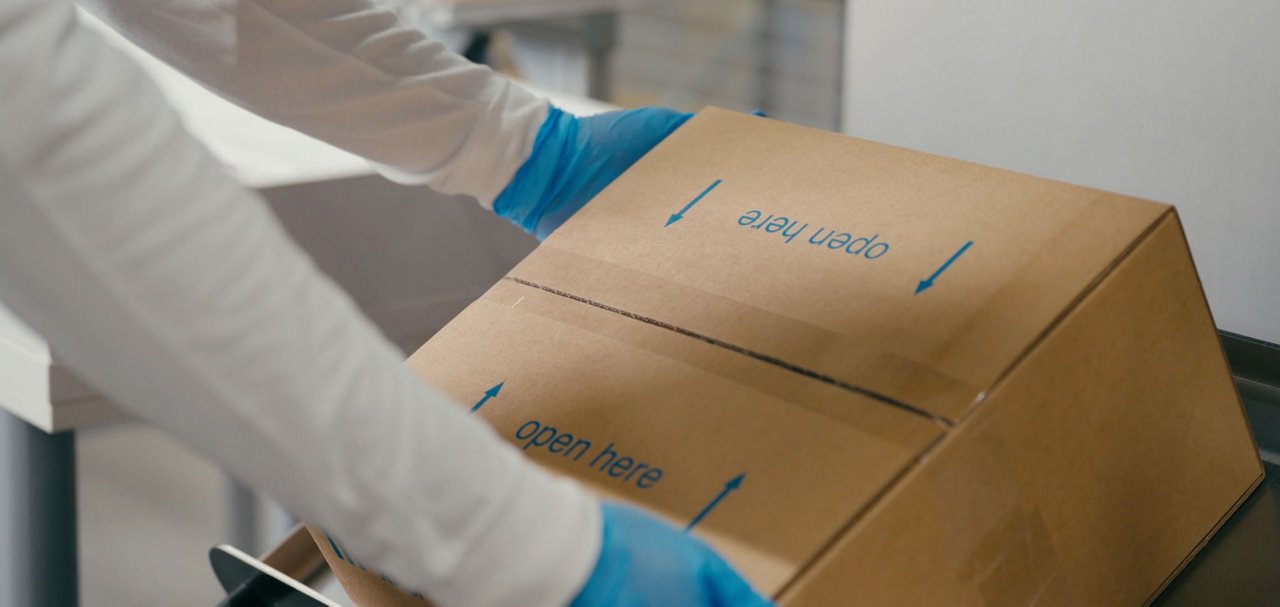 A person wearing a white protective garment and blue gloves handles a large cardboard box in a clean indoor setting.
