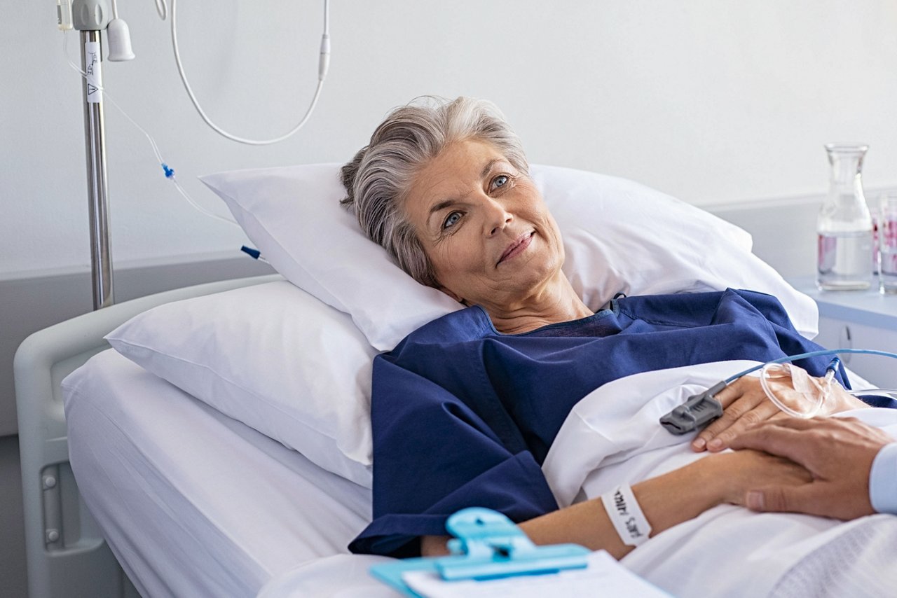 Patient lying in a hospital bed with an IV line and monitoring equipment while another person holds the patient’s hand.