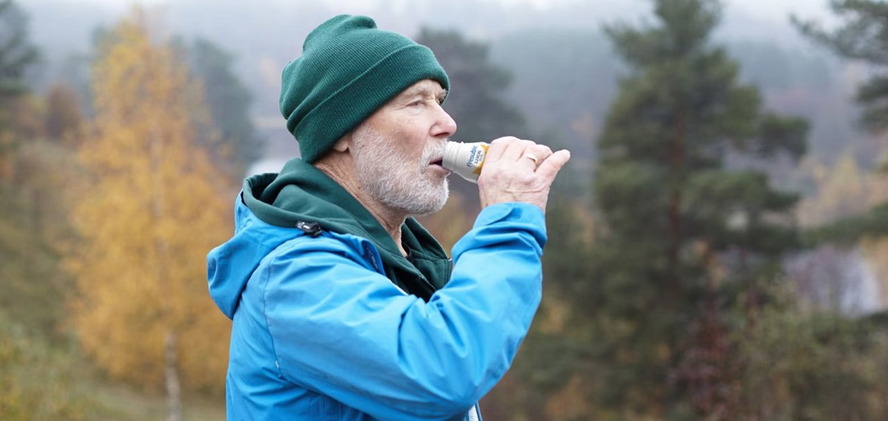 A person outdoors drinks a nutritional beverage while standing in a natural landscape.