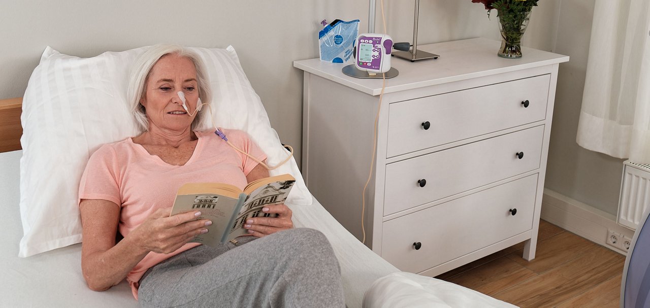 A person reclines in bed reading a book while connected to a nutrition delivery system in a home environment.