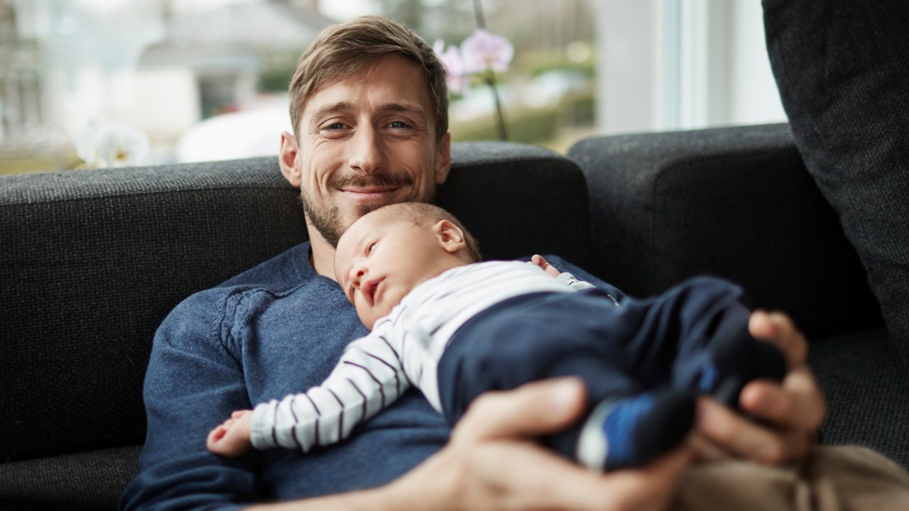 Person reclining on a sofa while holding an infant resting on their chest in a bright living room.