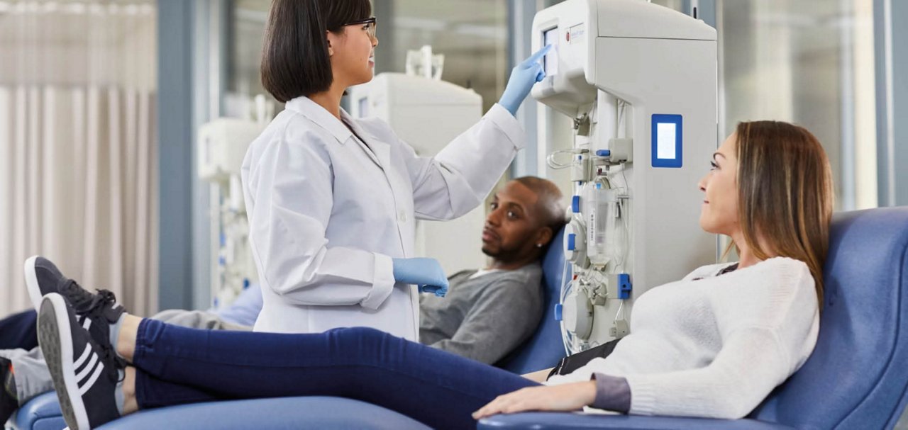 Individuals are seated in treatment chairs beside plasma collection equipment while a healthcare professional operates a touchscreen control panel in a clinical setting.
