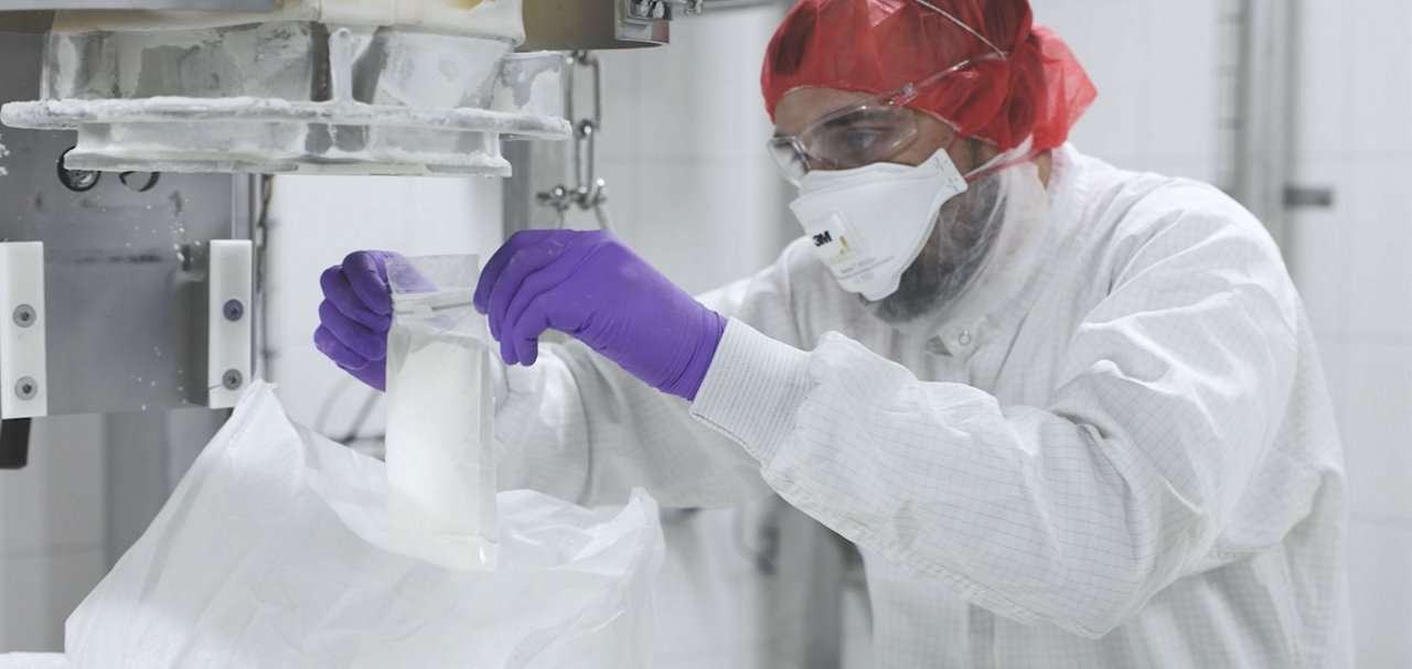 Person wearing protective clothing, safety glasses, a face mask and purple gloves handling a container beside production equipment