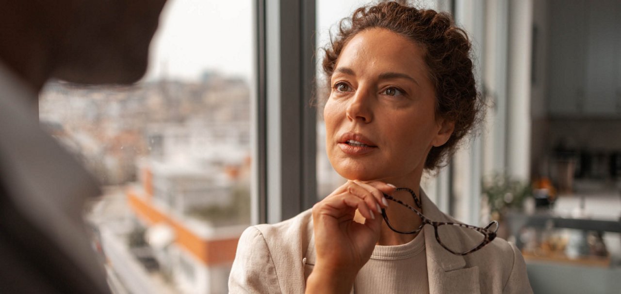 Two people having a conversation by a large office window, with one person holding eyeglasses