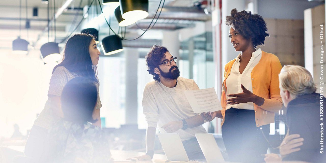 Business people looking at female colleague explaining documents while standing at conference table
