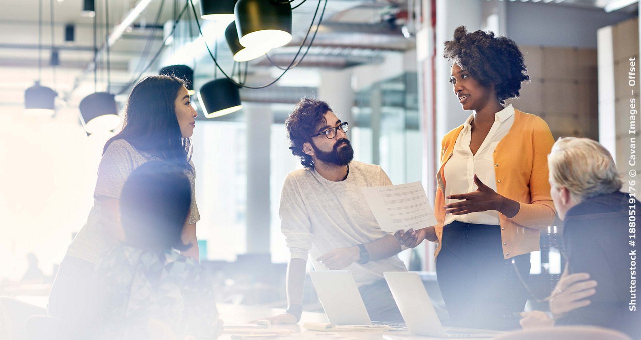 Business people looking at female colleague explaining documents while standing at conference table