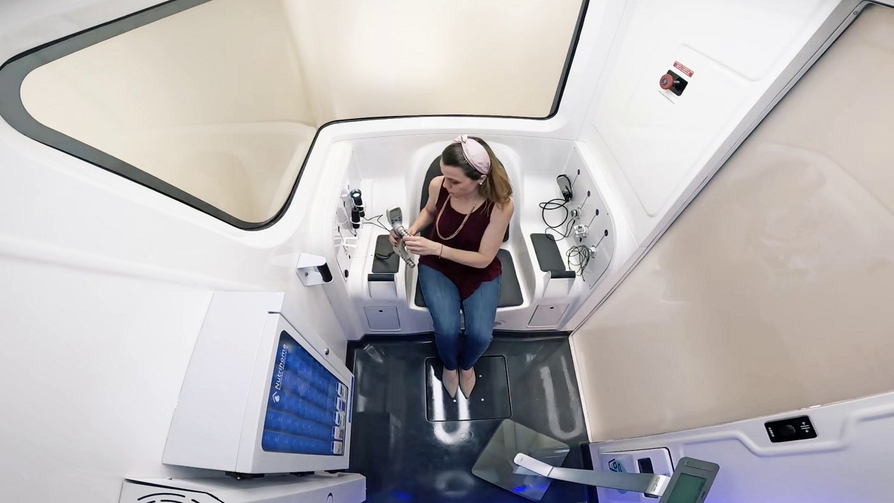 Individual seated inside a high-tech medical diagnostics pod, surrounded by monitoring equipment, screens, and cables suggesting a sterile, technology-driven environment for health assessment or clinical evaluation.