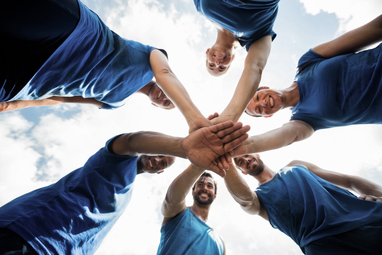 A group of people standing in a circle, all wearing blue shirts, with their hands stacked together in the center against a backdrop of a cloudy sky.