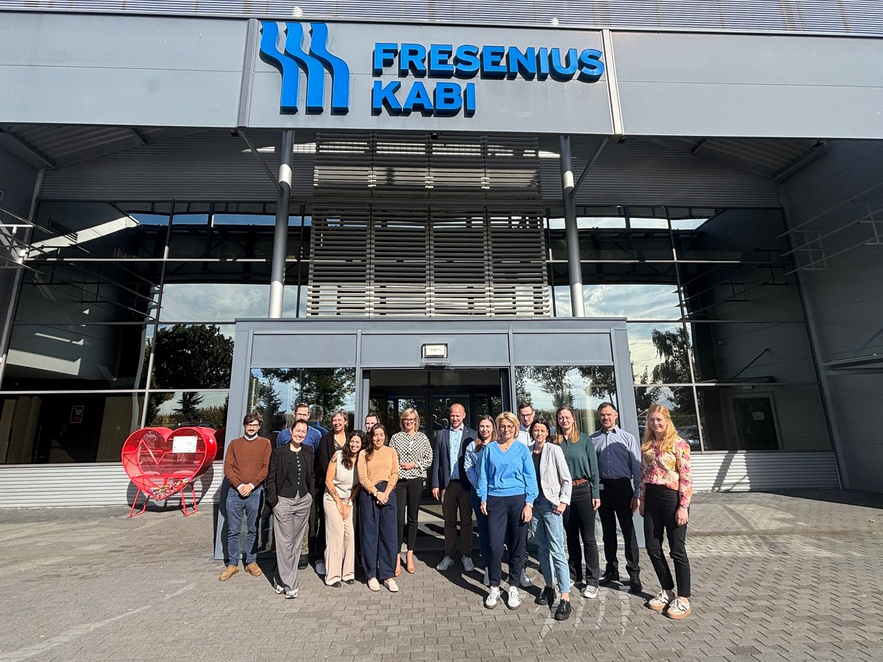 Group of people standing outside a Fresenius Kabi office building entrance. A large corporate sign is mounted above the entrance and the group is assembled in front of the facility.