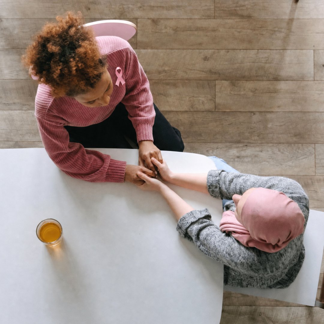 Two people holding hands across a table, with one wearing a pink breast cancer awareness ribbon.