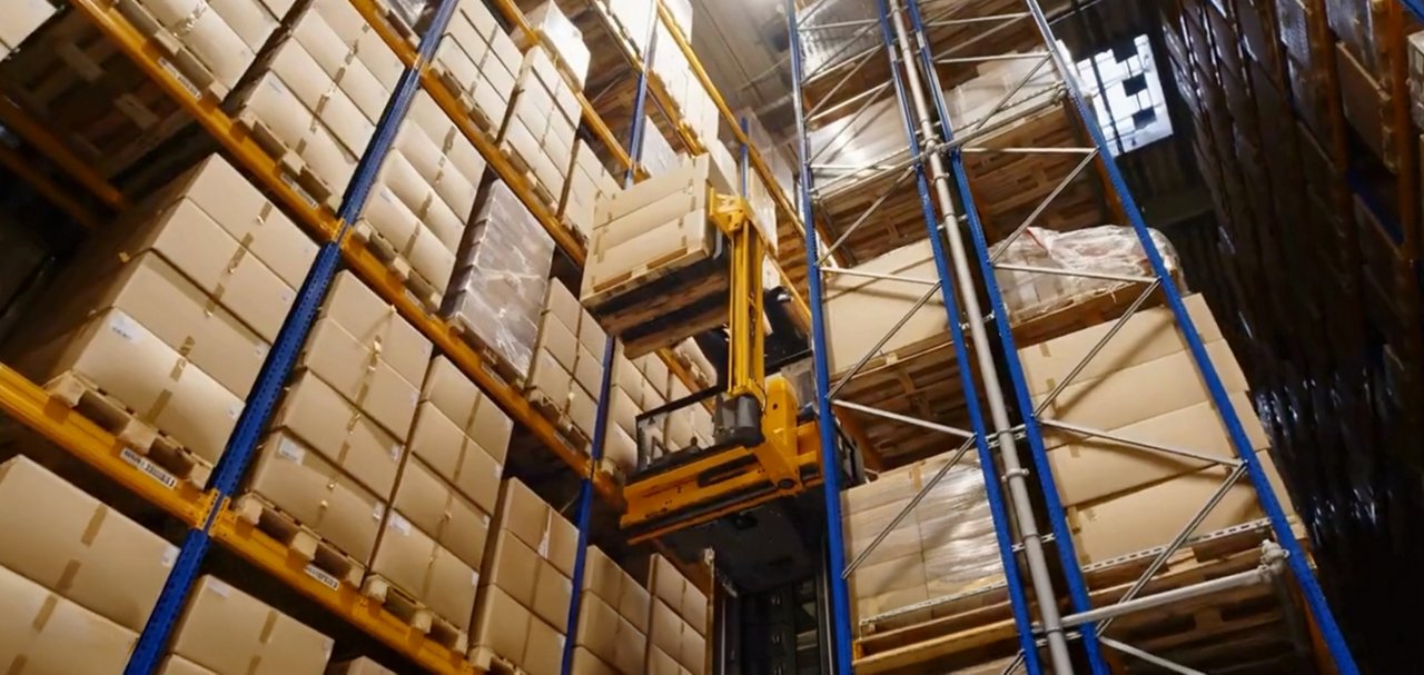 Material-handling equipment moving a pallet between tall warehouse racks filled with cardboard boxes.