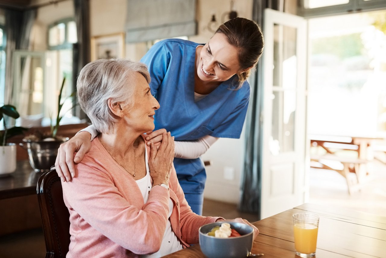 Healthcare, elderly woman with nurse with breakfast at her home and at the table in living room. Support or communication, caregiver and conversation with medical person with senior or old female.